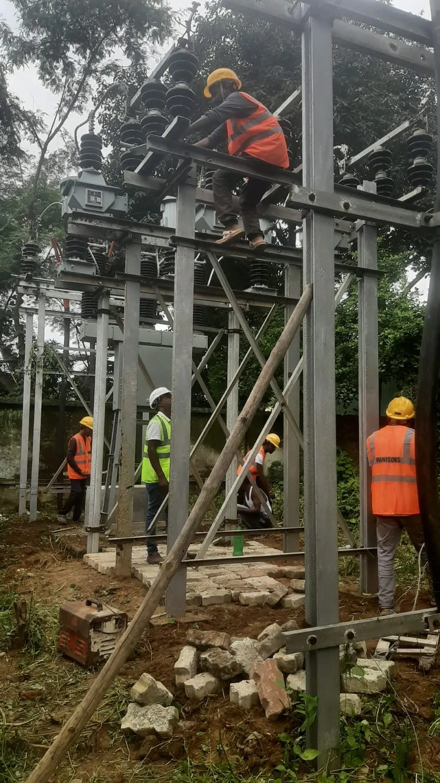 Lineman working at height on an isolator gantry crossarm while four crew work at ground level below